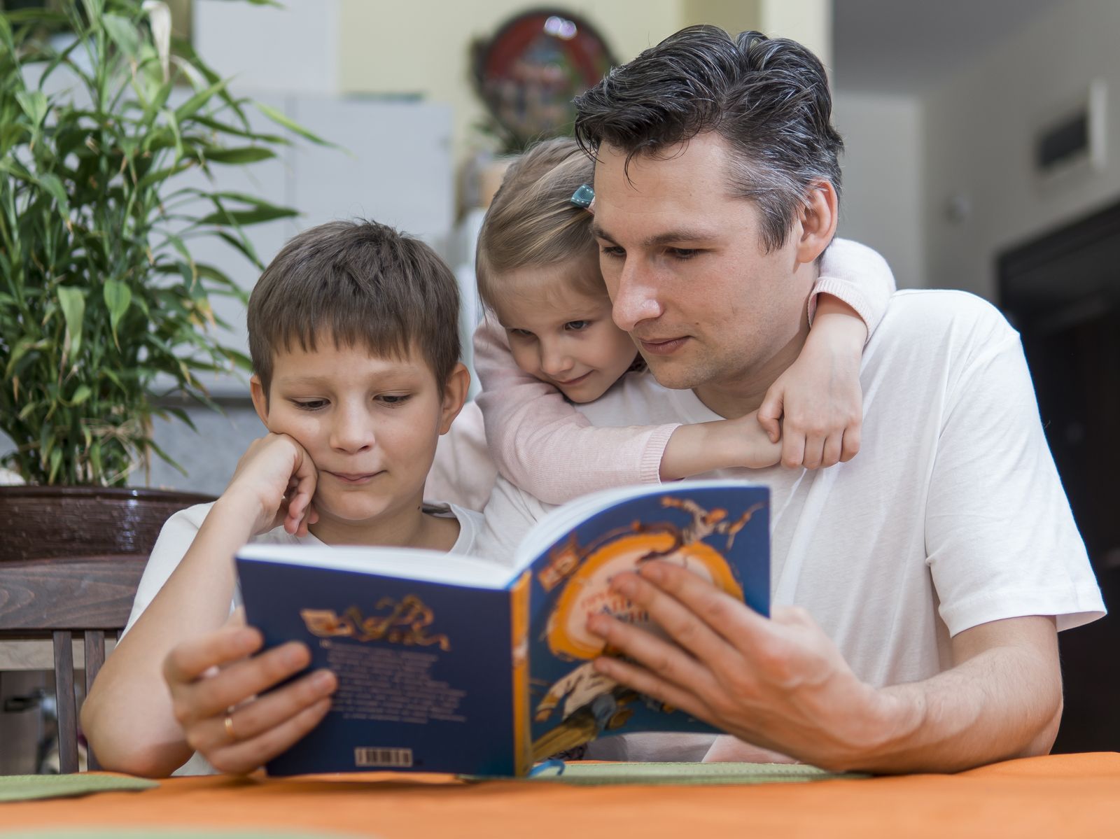 Father reading a book with his two children at the dining table